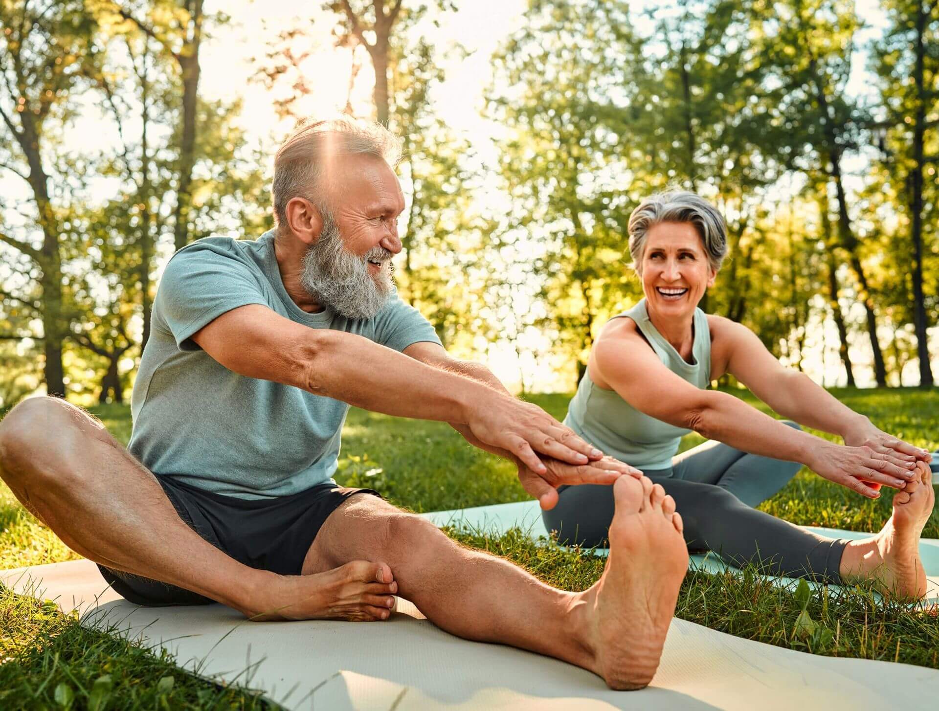 A smiling senior man and woman in athletic wear sitting on yoga mats in a sunlit park, performing seated forward-fold stretches together.